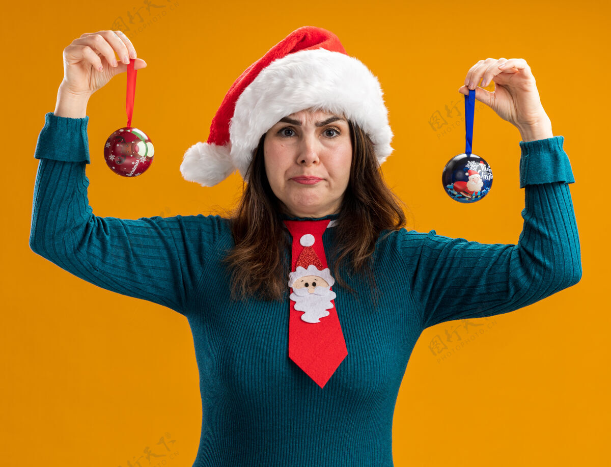 玻璃愤怒的成年白人妇女戴着圣诞帽 打着圣诞领带 手里拿着玻璃球饰物 孤立地放在橙色背景上 还有复制空间成人帽子装饰品