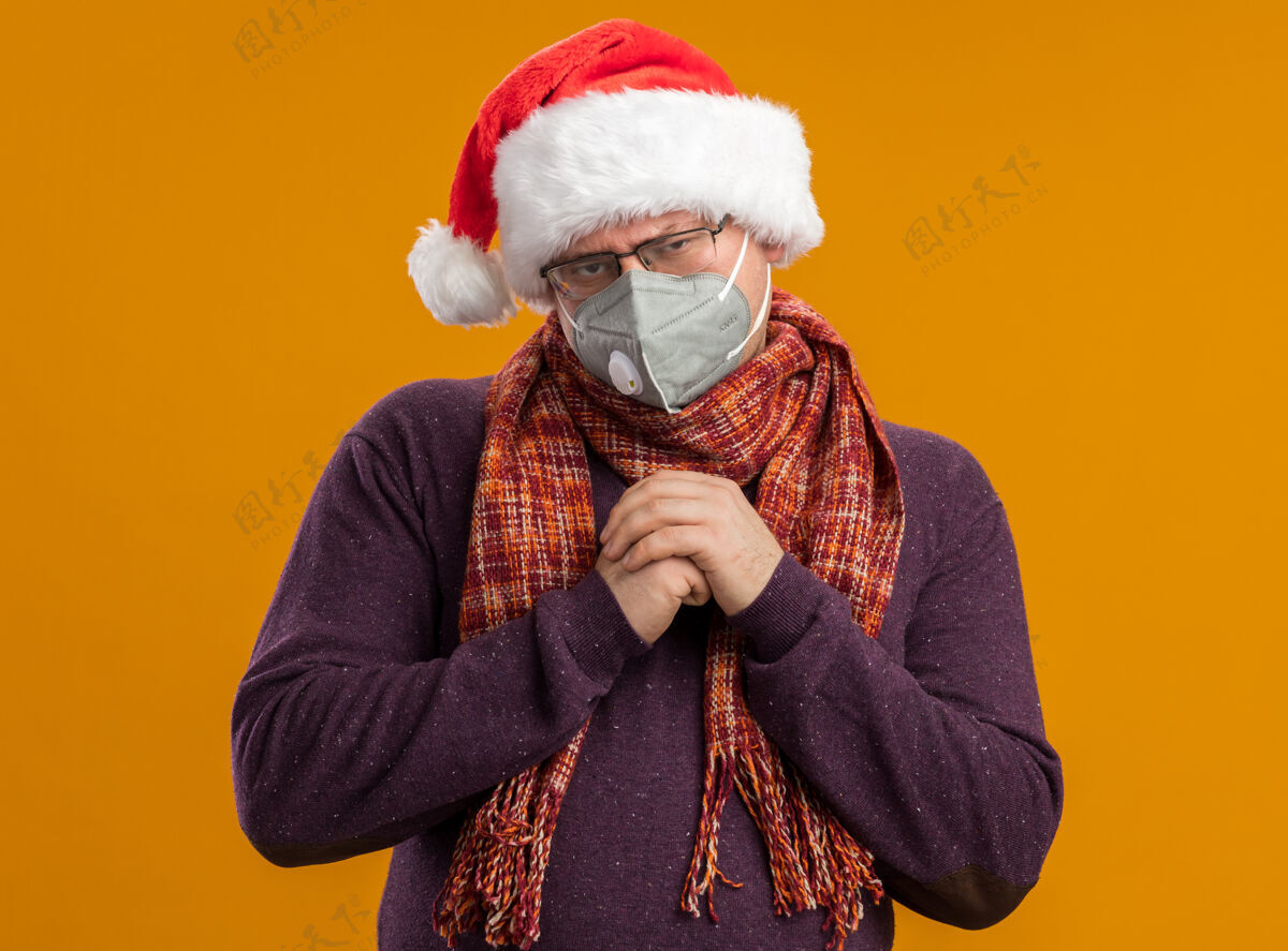 围巾狡猾的成年男子戴着防护面具眼镜 戴着圣诞老人帽 脖子上围着围巾 手放在一起看着橙色背景上孤立的摄像机脖子成人保护