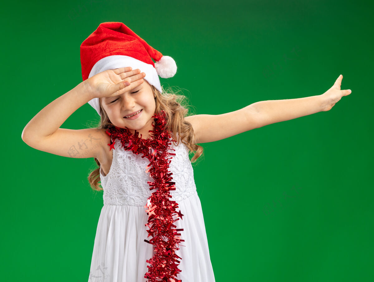 未放小女孩戴着圣诞帽 脖子上戴着花环 手放在额头上 手指放在一边 在绿色背景上隔离 留有复印空间帽子侧面小