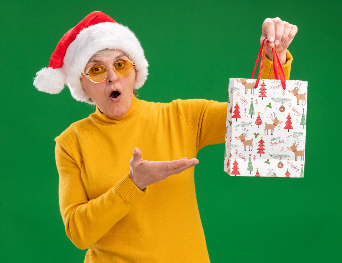 指着一位戴着太阳眼镜 戴着圣诞老人帽子的惊讶的老妇人拿着一个纸礼包 这个纸礼包被隔离在绿色的背景上 还有复印空间帽子包拿着