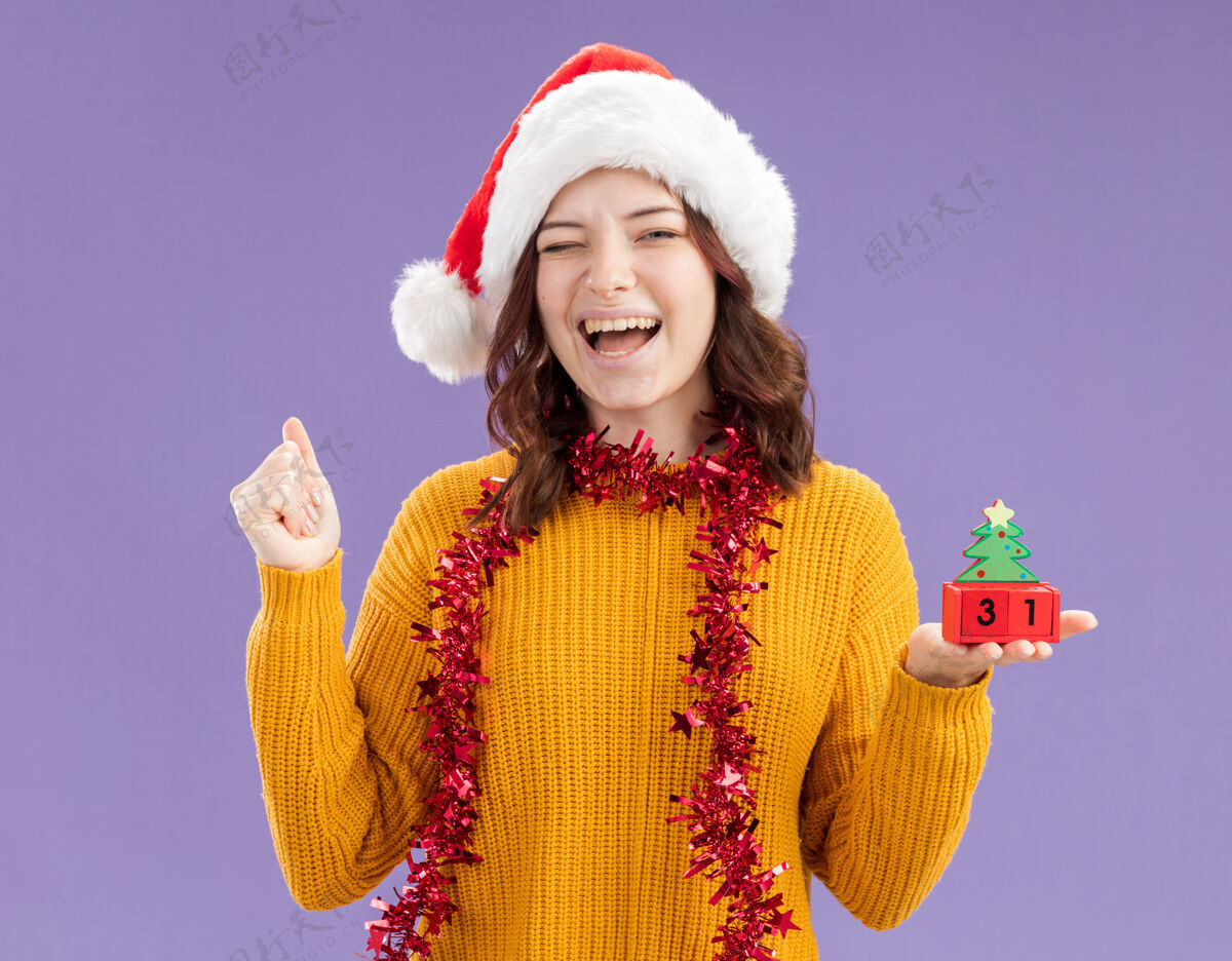 圣诞快乐的年轻斯拉夫女孩戴着圣诞帽 脖子上戴着花环 眨着眼睛 拿着圣诞树上的装饰物 在紫色的背景上与复制空间隔离开来圣诞老人女孩装饰品