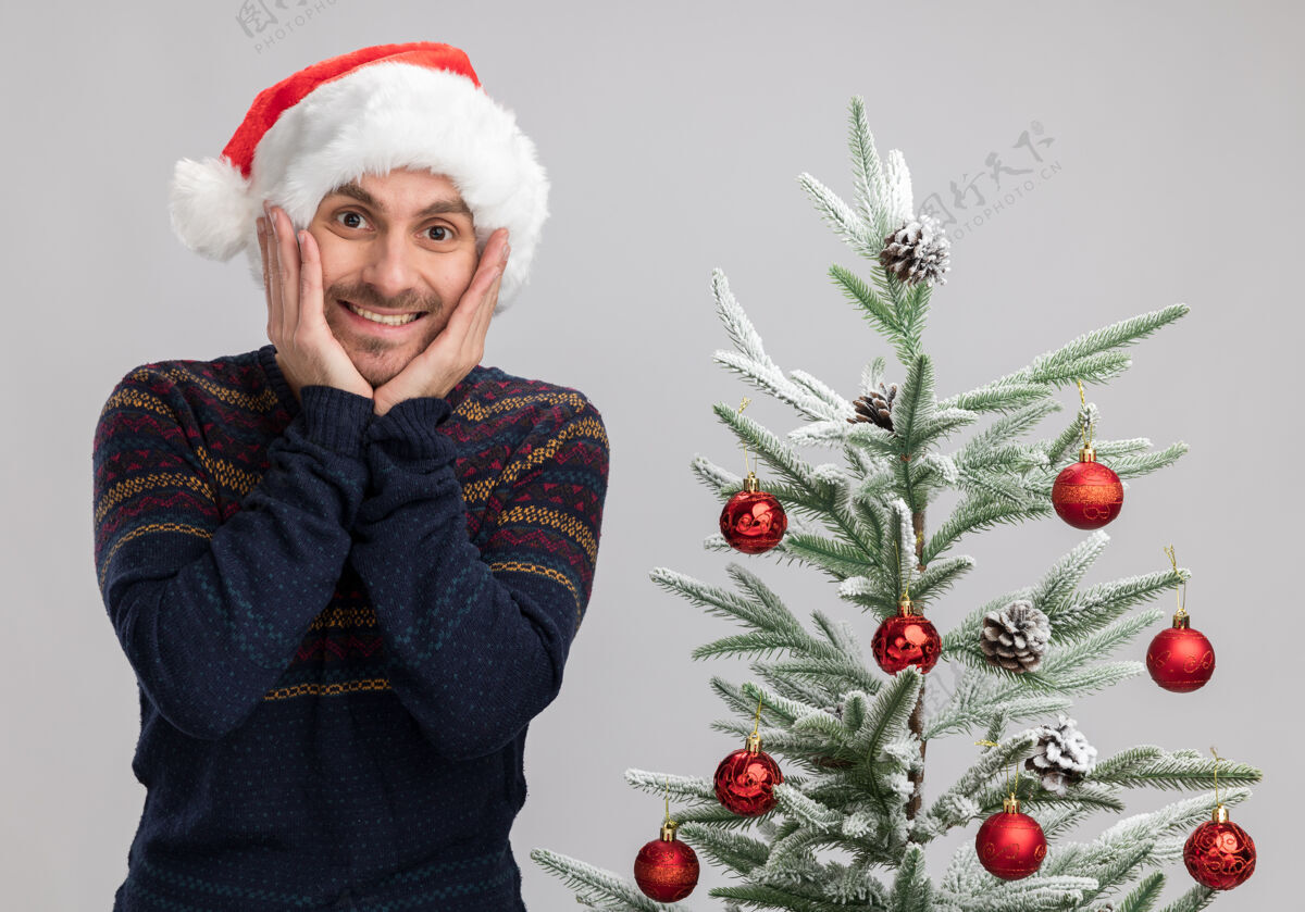 年轻兴奋的白人年轻人戴着圣诞帽 站在圣诞树旁 手放在隔离的白色墙上保持靠近站着