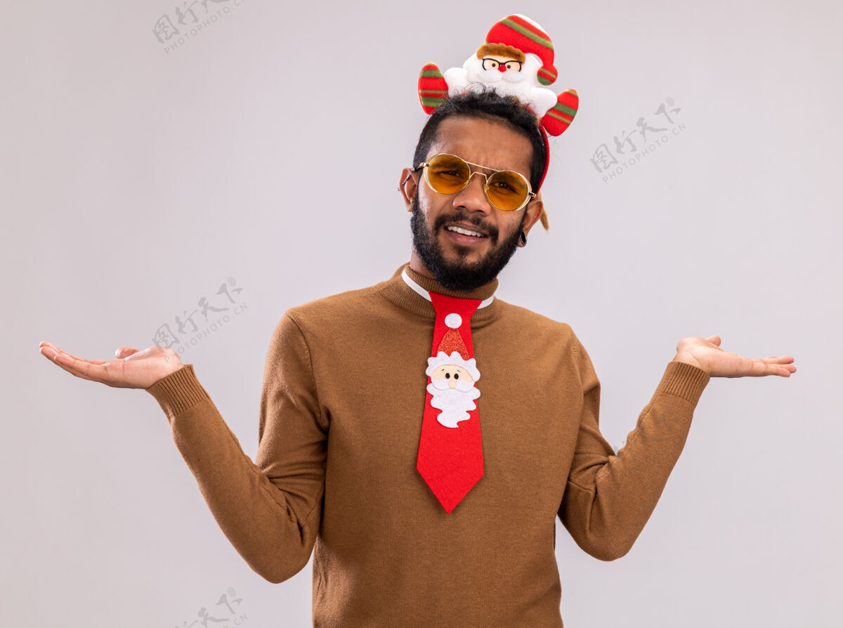 美国人一个身穿棕色毛衣 头戴圣诞老人项圈 打着有趣的红色领带的非裔美国人站在白色背景下困惑而不高兴地看着摄像机站着边缘头