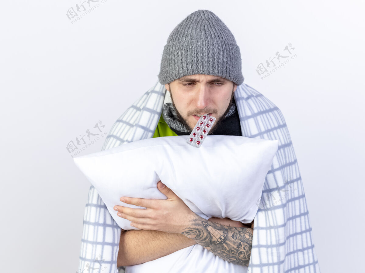 医学苍白的年轻白种人 戴着格子布裹着的冬季帽子 手里拿着一包带牙齿的胶囊 抱着一个隔离在白色背景上的枕头帽子男人空间
