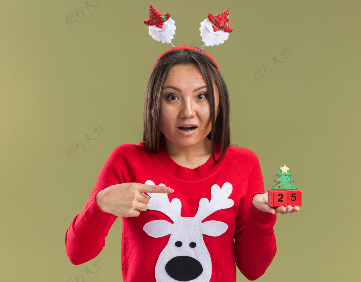 亚洲人困惑的年轻亚洲女孩戴着圣诞发箍 指着橄榄绿背景上孤立的圣诞玩具箍点女孩