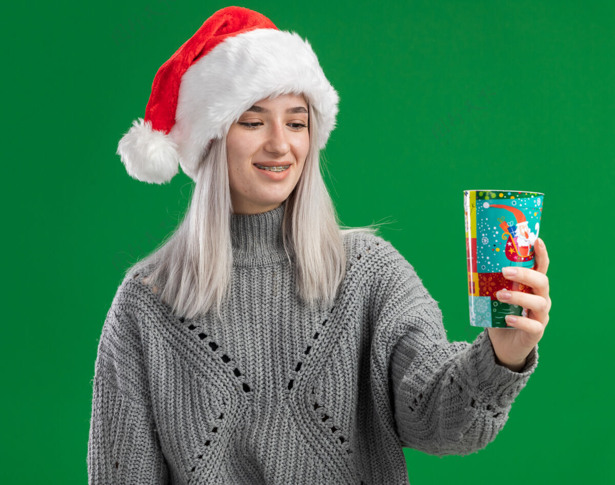圣诞老人年轻的金发女郎穿着冬衣 戴着圣诞帽 手里拿着五颜六色的纸杯 站在绿色的背景下 面带微笑地看着纸杯年轻脸毛衣