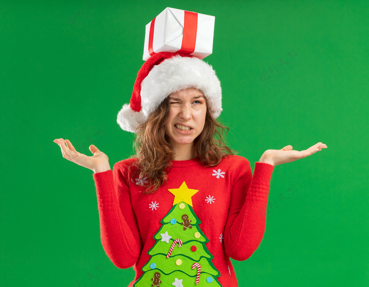 不高兴身穿红色圣诞毛衣 头戴圣诞老人帽 头上戴着礼物的年轻女子 神情迷茫 不高兴地张开双臂站在绿色背景上站立帽子头