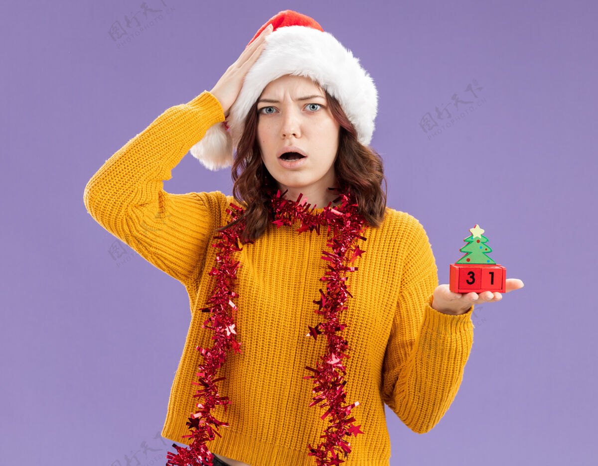 装饰品震惊的年轻斯拉夫女孩戴着圣诞帽 脖子上戴着花环 手放在头上 手拿着圣诞树饰物 背景是紫色的 有复制空间举行花环树