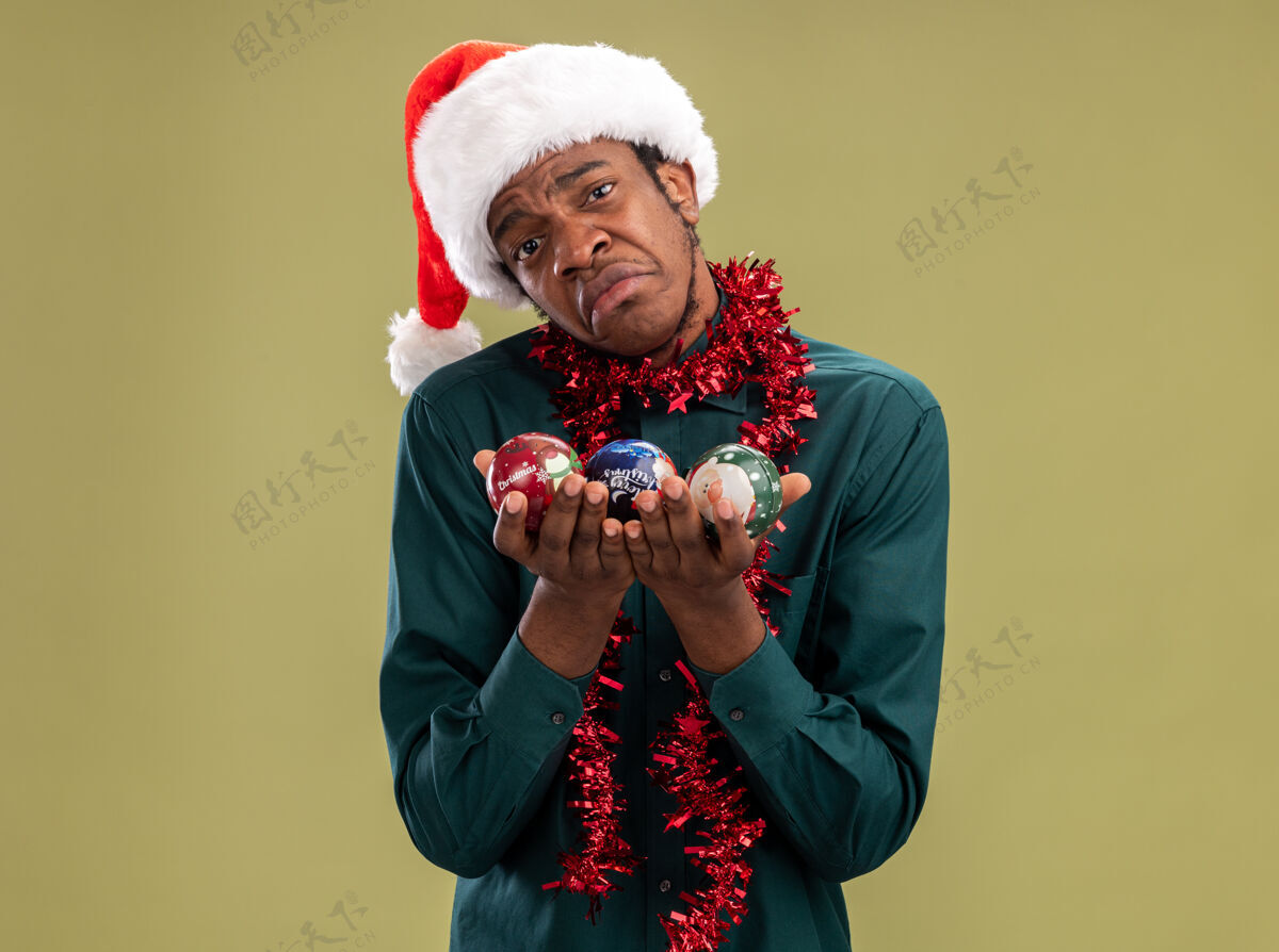 美国人心烦意乱的非洲裔美国人戴着圣诞帽 戴着花环 手里拿着圣诞球 站在绿色的背景上 愁眉苦脸地撅着嘴唇看着摄像机抱着表情站着