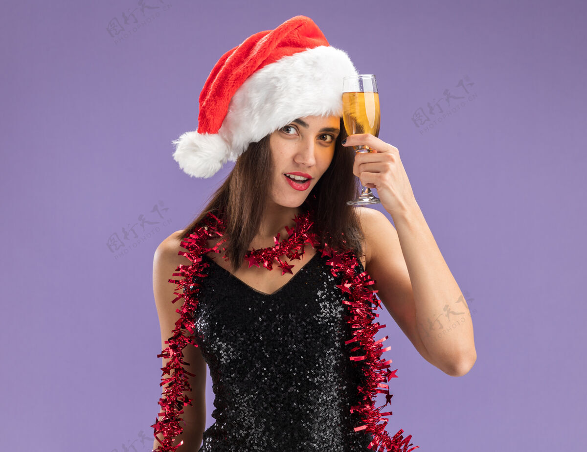 女孩给人印象深刻的年轻漂亮女孩戴着圣诞帽 脖子上戴着花环 额头上拿着一杯香槟酒 紫色的背景隔离开来印象深刻年轻圣诞节