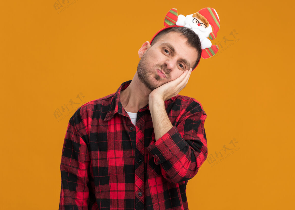 保持无聊的白人年轻人戴着圣诞老人的头带看着相机手贴在脸上 噘着嘴唇 在橙色的背景下与复制空间隔离相机脸快乐