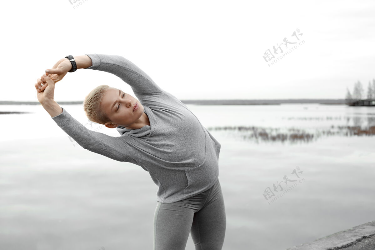 健身积极的生活方式 瑜伽 健身和运动理念户外视野：强壮柔韧的年轻白种女性跑步者伸展双臂 向一侧弯腰 温暖身体 早晨沿着河岸跑步活动天空活动