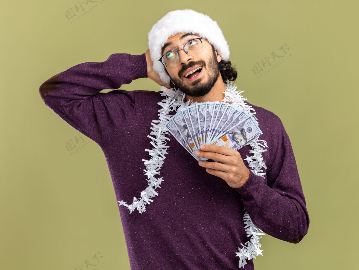 花环面带微笑的年轻帅哥戴着圣诞帽 脖子上戴着花环 手里拿着现金 手放在头上 背景是橄榄绿微笑小伙子放
