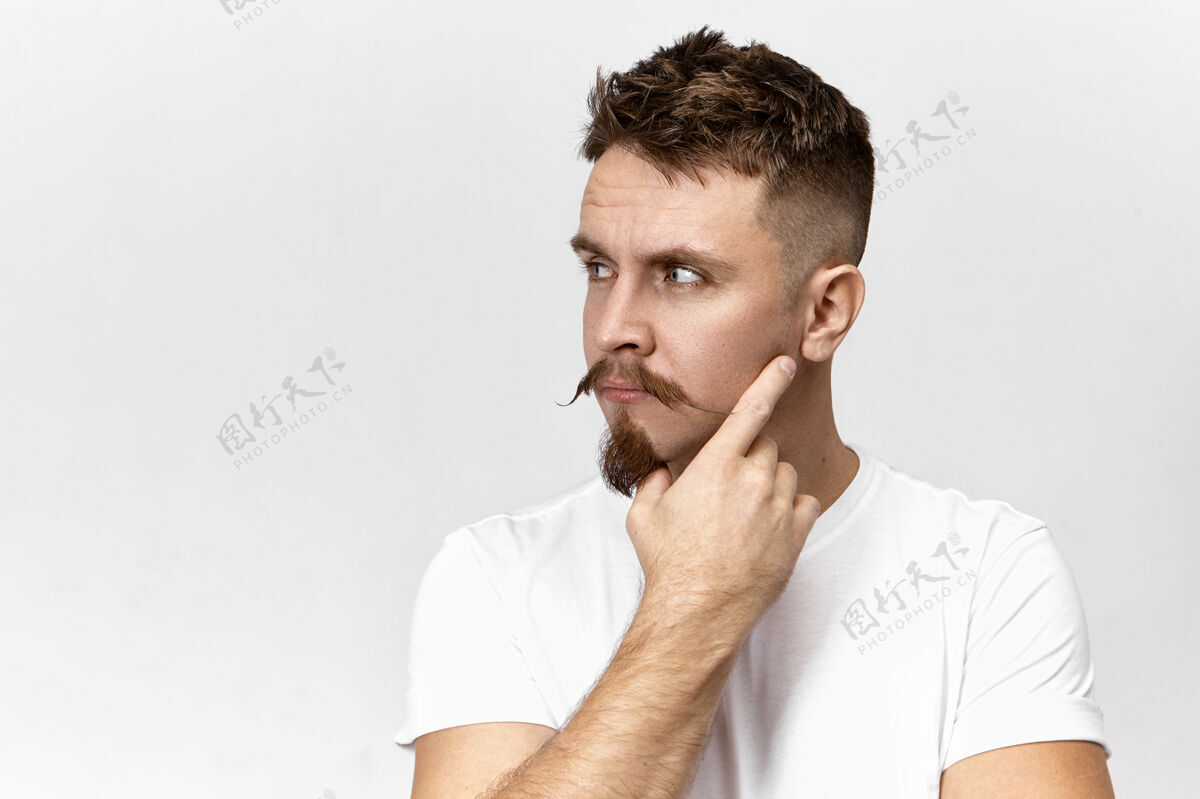 年轻迷人时尚的年轻留胡子的家伙 留着胡子 想着什么 手放在脸上 在空白的工作室背景墙上摆姿势 留着广告信息的复制空间胡须时尚脸