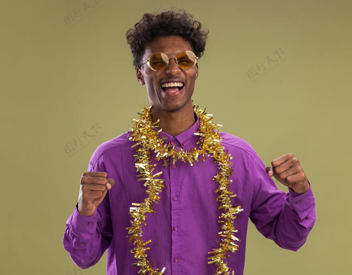 是的快乐的非洲裔美国年轻人戴着眼镜 脖子上戴着金箔花环 看着相机做着“是”的手势 在橄榄绿的背景下孤立着周围金属片做