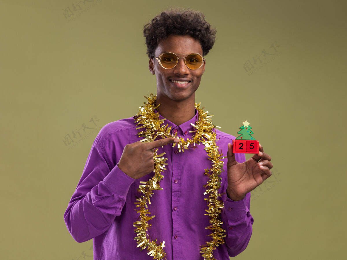 举行带着微笑的非洲裔美国年轻人戴着眼镜 脖子上戴着金属丝花环 拿着并指着圣诞树玩具和日期 看着橄榄绿背景上孤立的相机花环树眼镜