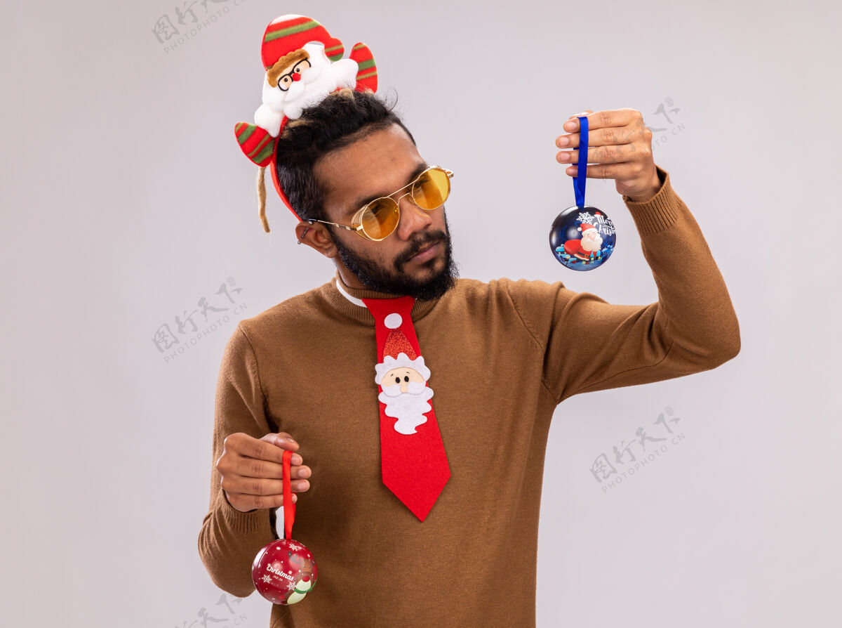 圣诞节一个非洲裔美国人 穿着棕色毛衣 头戴圣诞老人圈 打着有趣的红色领带 手里拿着圣诞球 站在白色的背景上 看起来很困惑 有疑问轮辋站立举行
