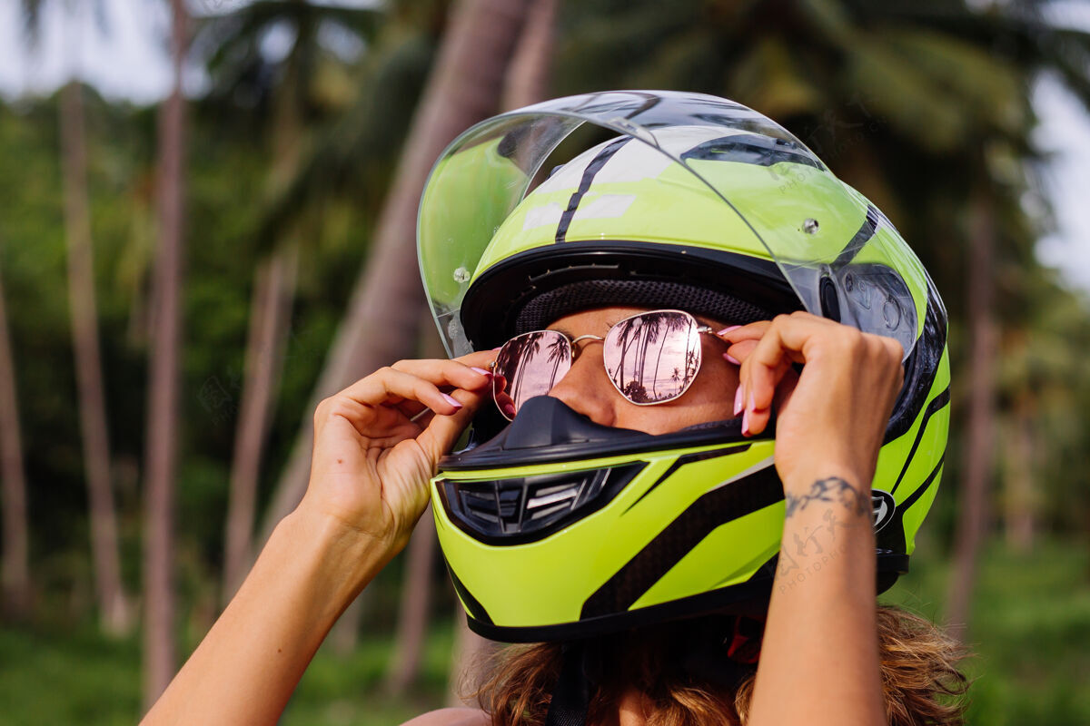 骑手在棕榈树下热带田野的丛林中 穿着黄绿色摩托车头盔和彩色浅夏装的美丽骑手妇女的肖像自行车热带度假