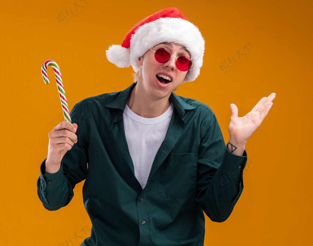 印象深刻令人印象深刻的年轻金发男子戴着圣诞帽 戴着圣诞眼镜 拿着圣诞甜手杖 看着相机 显示出一只孤立在橙色背景下的空手手杖圣诞老人眼镜