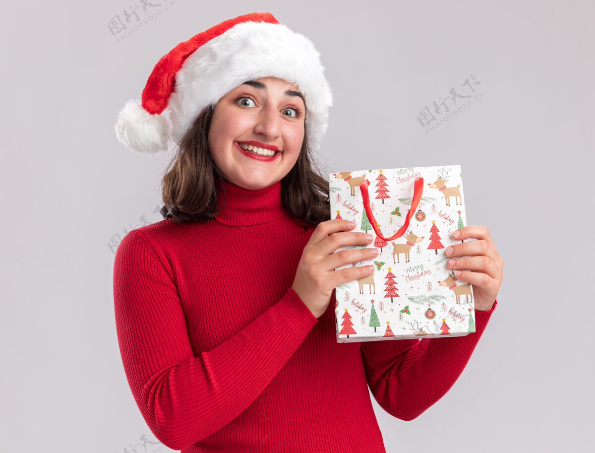 女孩穿着红色毛衣 戴着圣诞帽 手里拿着彩色纸袋 手里拿着圣诞礼物的快乐小女孩站在白色的背景下 面带微笑地看着相机圣诞节包脸