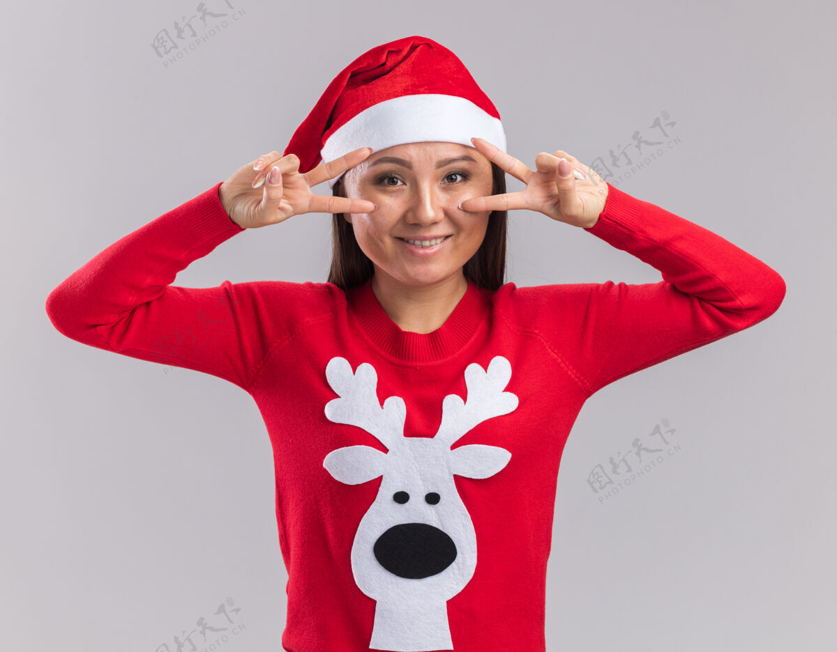 帽子笑容满面的年轻亚洲女孩戴着圣诞帽 穿着毛衣 在白色背景上显示出与世隔绝的和平姿态亚洲人年轻微笑