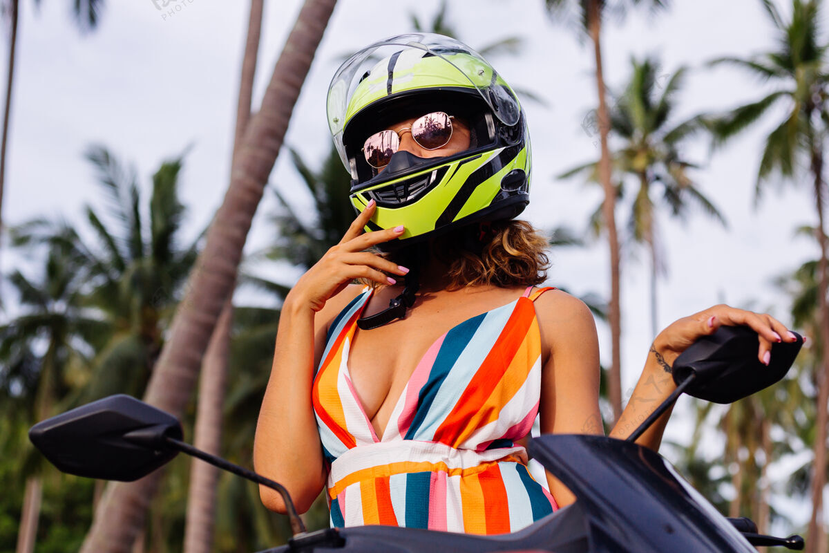 租赁在棕榈树下热带田野的丛林中 穿着黄绿色摩托车头盔和彩色浅夏装的美丽骑手妇女的肖像肖像手掌户外