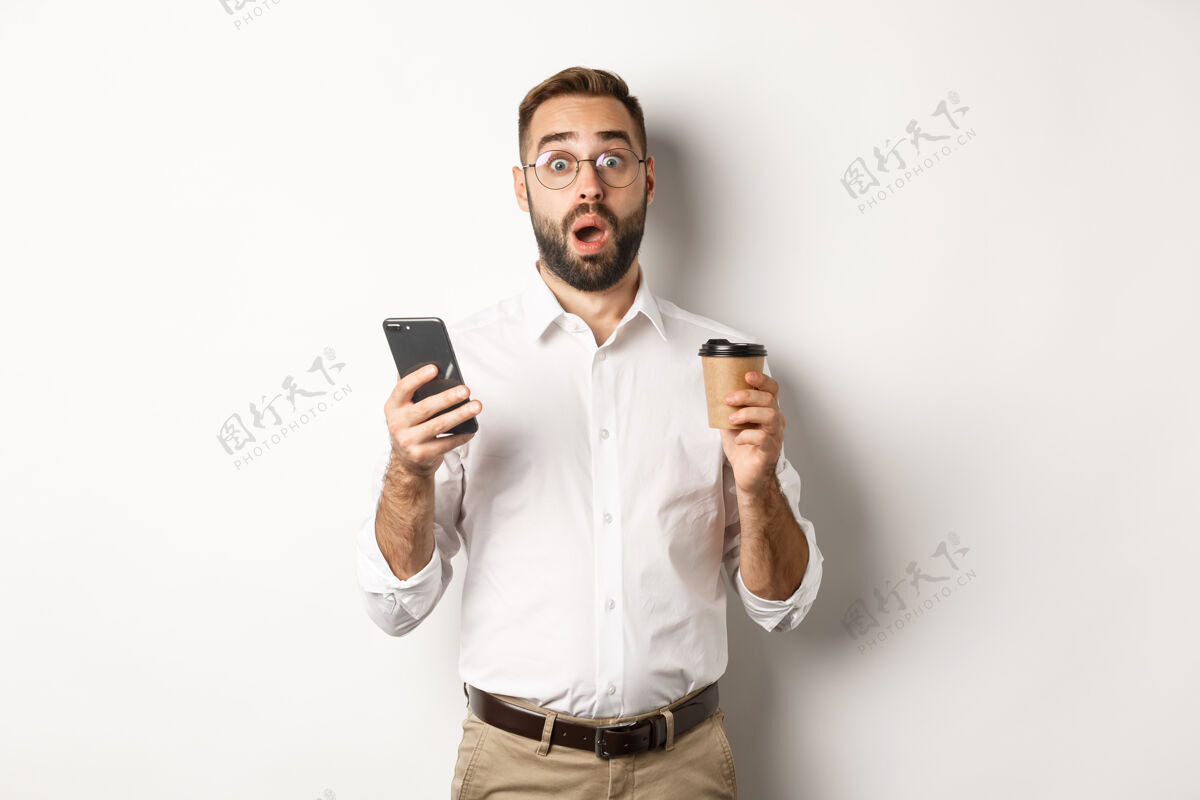 互联网帅哥喝着咖啡 对手机上的留言反应惊讶 站着胡须印象深刻智能手机