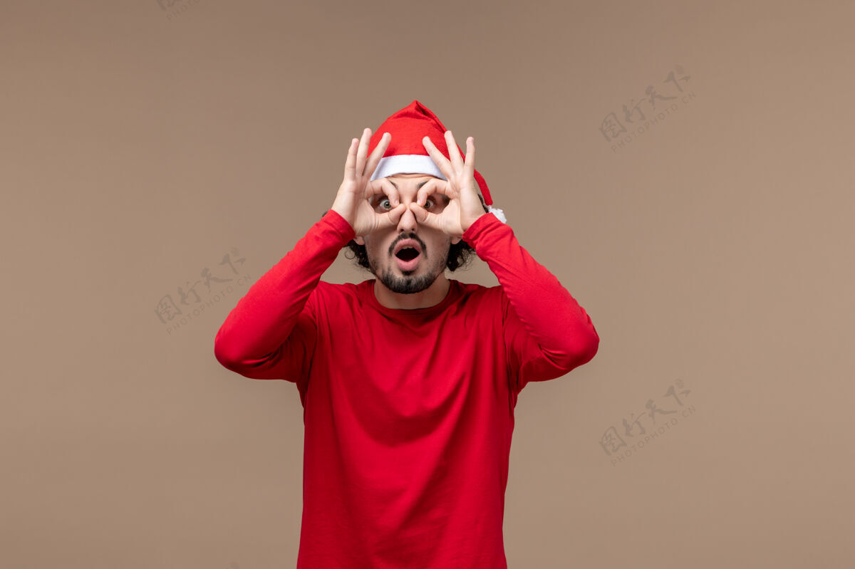 喜剧演员前视图年轻的男性透过手指看棕色背景的情感圣诞假期封面衣服人