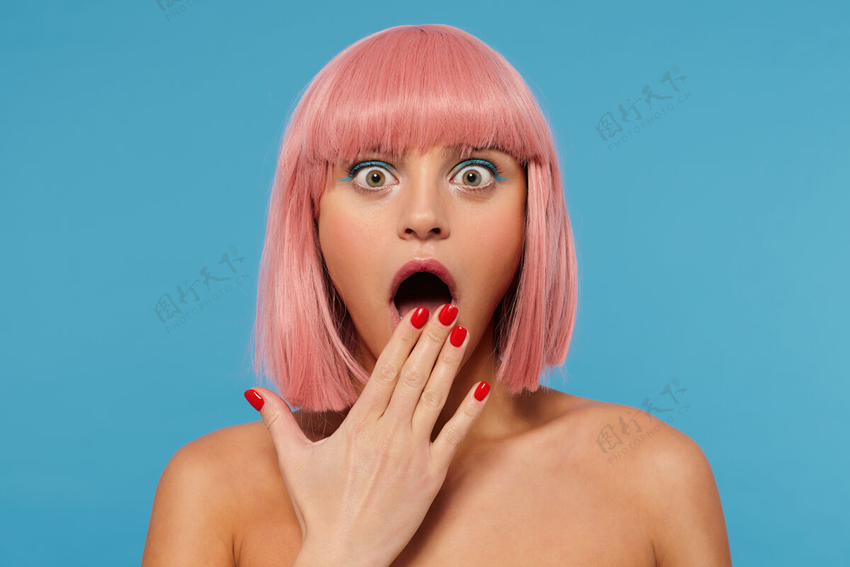 发型震惊的绿眼睛年轻的粉红色头发的女性惊奇地看着镜头与圆眼睛 并涵盖了她张开的嘴与提高手掌 在蓝色的背景摆姿势优雅惊讶姿势
