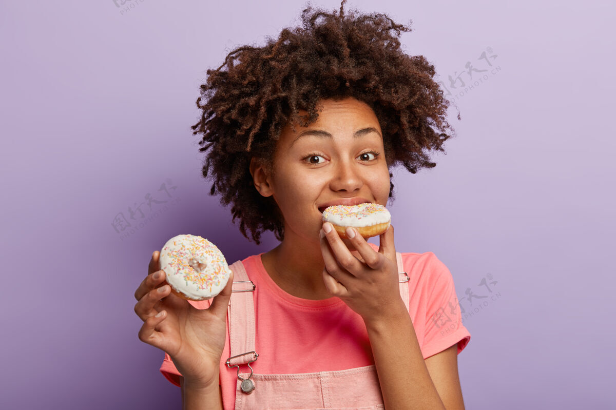 肖像饥肠辘辘的非洲女人咬着鲜亮的甜甜圈 有不健康的营养 无法想象没有甜点的生活 卷发 不节食 孤立在紫色的墙上非洲零食青少年