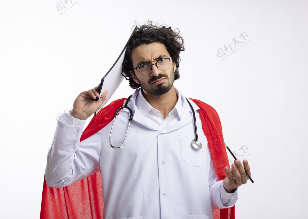 超级英雄年轻的白人超级英雄戴着眼镜 身穿医生制服 披着红色斗篷 脖子上戴着听诊器 把剪贴板放在头上 把铅笔放在白墙上放置制服剪贴板