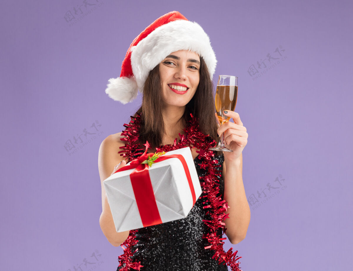 抱着微笑着的年轻漂亮女孩戴着圣诞帽 脖子上戴着花环 手里拿着一个礼盒 紫色背景上隔着一杯香槟脖子花环圣诞节