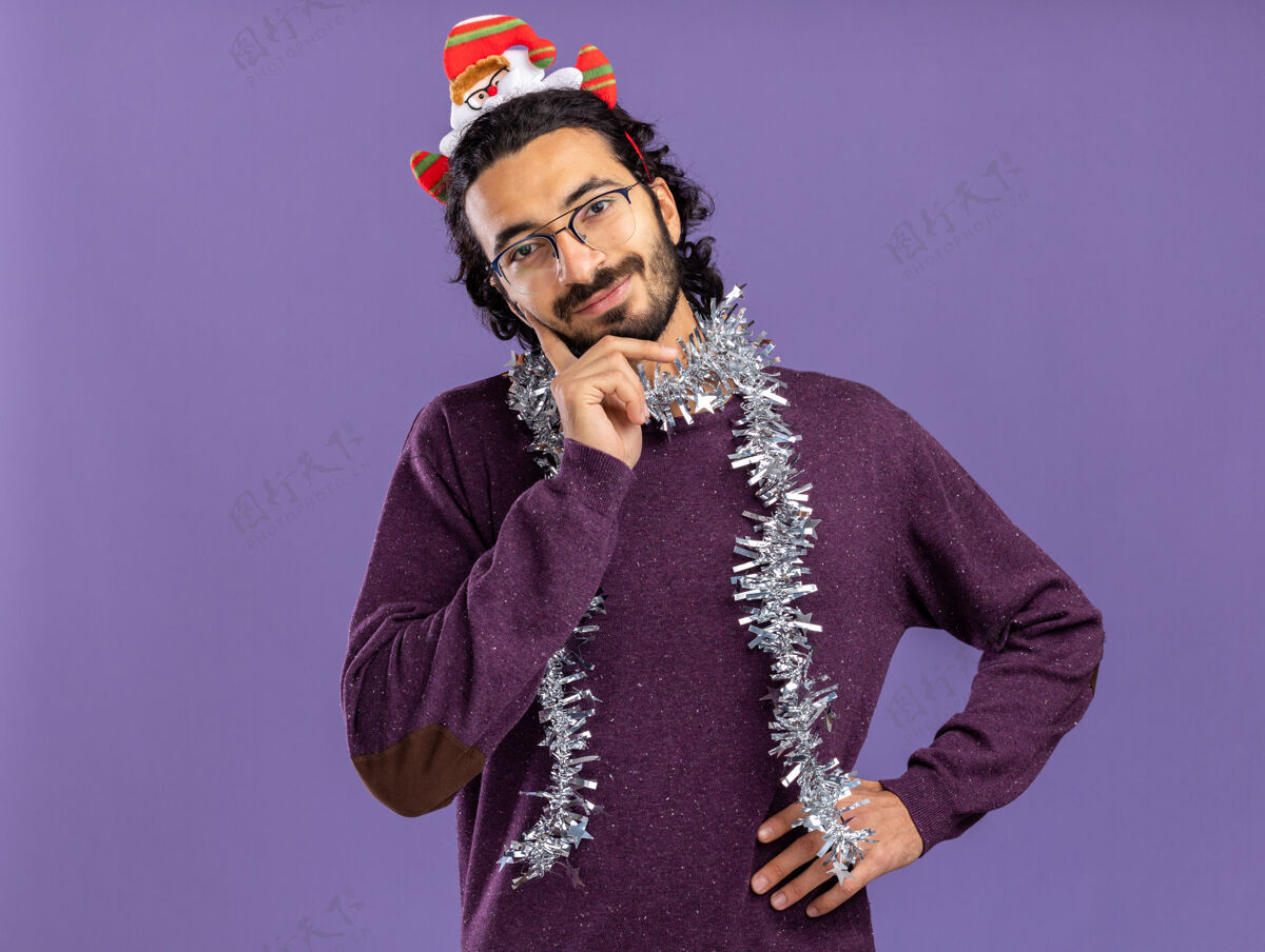 花环高兴的年轻帅哥戴着圣诞发箍 脖子上戴着花环 手放在脸颊上 臀部隔离在蓝色背景上小伙子高兴箍