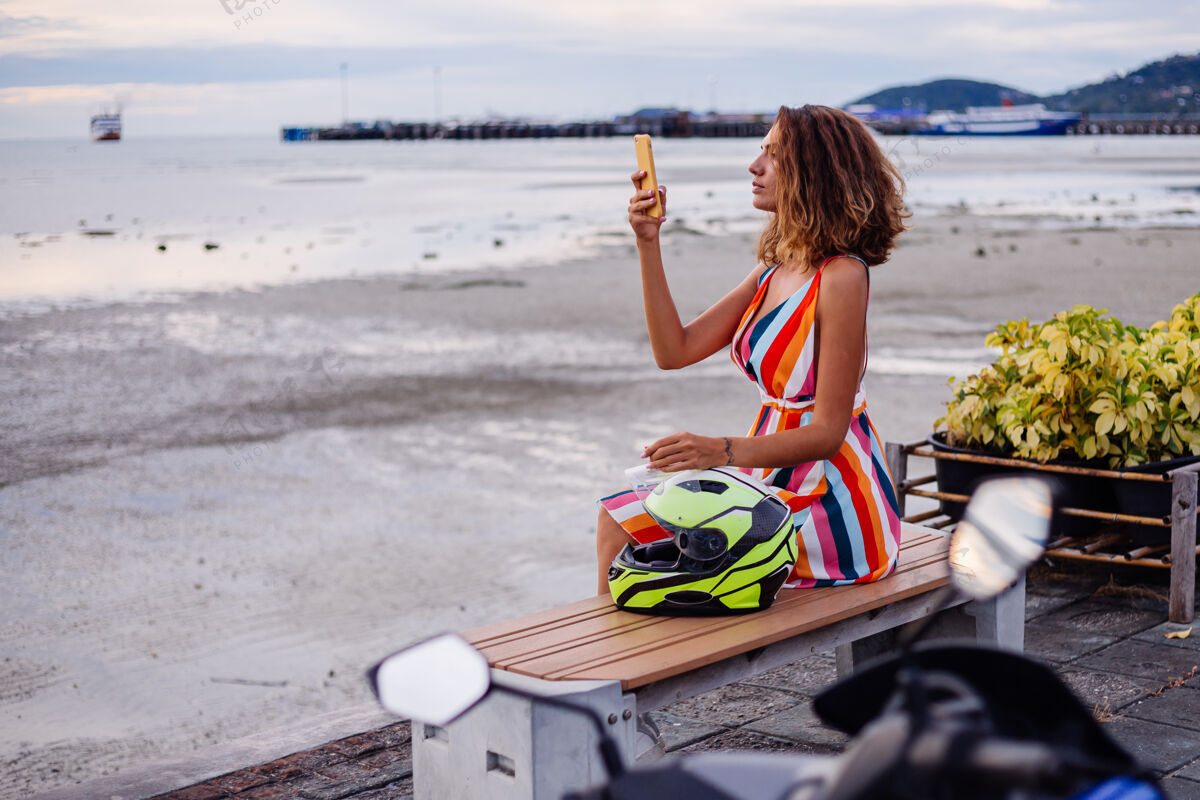 车辆快乐的高加索骑摩托车的女人 穿着五颜六色的夏装 戴着摩托车头盔度假热带旅游自行车