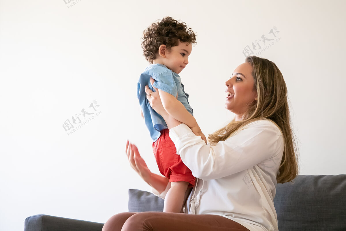 倾听长发的母亲抱着小男孩 和儿子聊天 坐在沙发上严肃的蹒跚学步的孩子站在妈妈的膝盖上 仔细地听她说话家庭时间 母亲和周末的概念儿子卷发膝盖