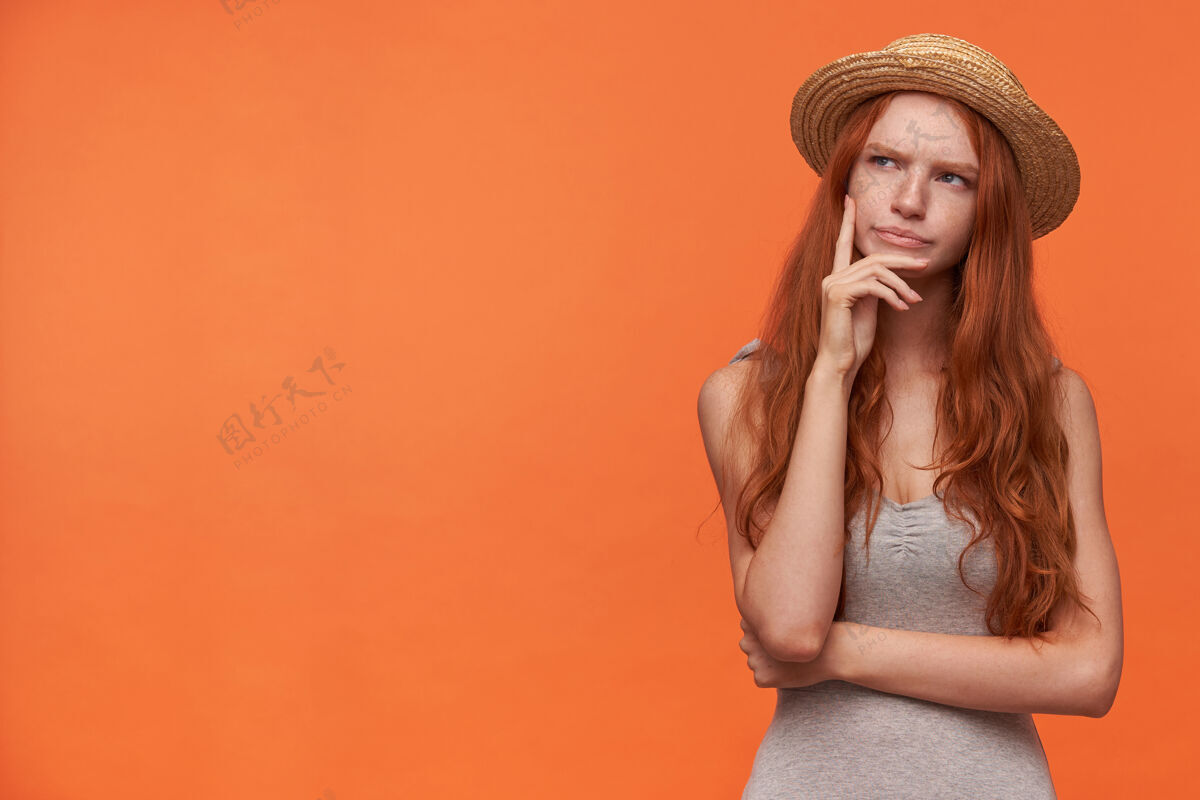 稻草摄影棚照片中的年轻女性 卷曲的狐狸尾巴 手放在下巴上 眉头紧锁 在橙色背景下穿着休闲服摆姿势下巴漂亮自然