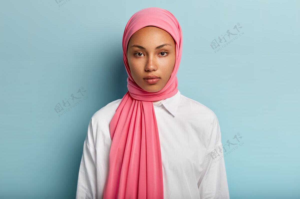 肖像表情冷静严肃的阿拉伯女人 皮肤黝黑 裹着粉色面纱 穿着白色衬衫 没有化妆 自然美丽 蓝墙之上的模特穆斯林女士特写镜头夹克凝视风格