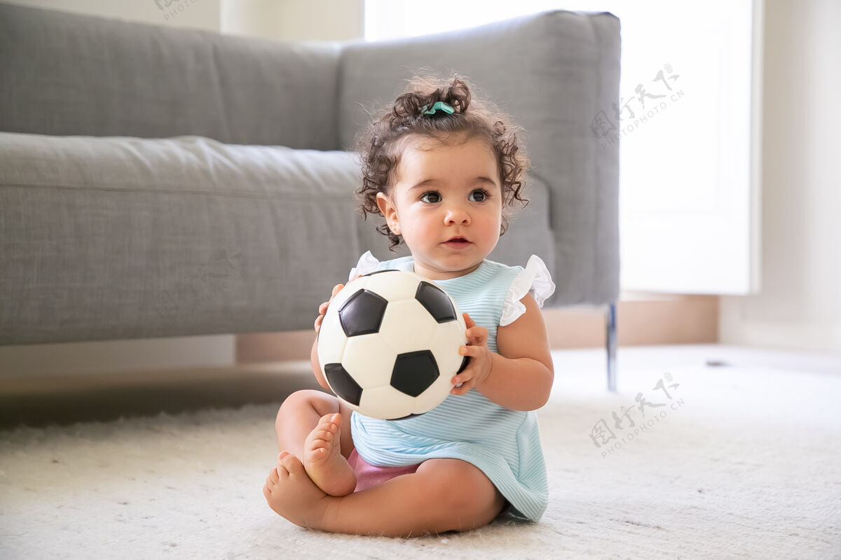 可爱可爱的黑发女婴 穿着淡蓝色的衣服 坐在家里的地板上 看着别处 踢足球家里的孩子和童年的概念卷发地毯孩子