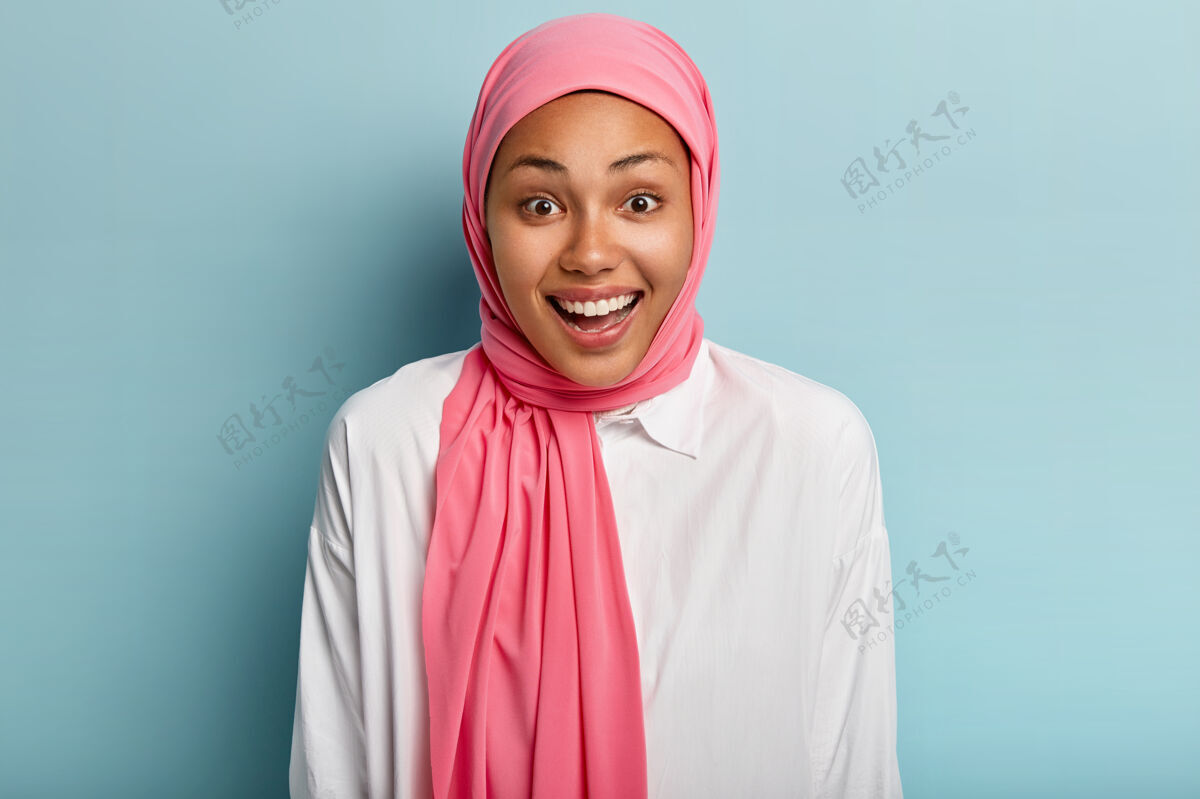 信仰喜怒无常的妇女欢笑 有愉快的表情 表达真挚的感情 戴着粉红色的头饰 露出白色的牙齿 隔着蓝色的墙壁人类情感的概念过度运动凝视头巾