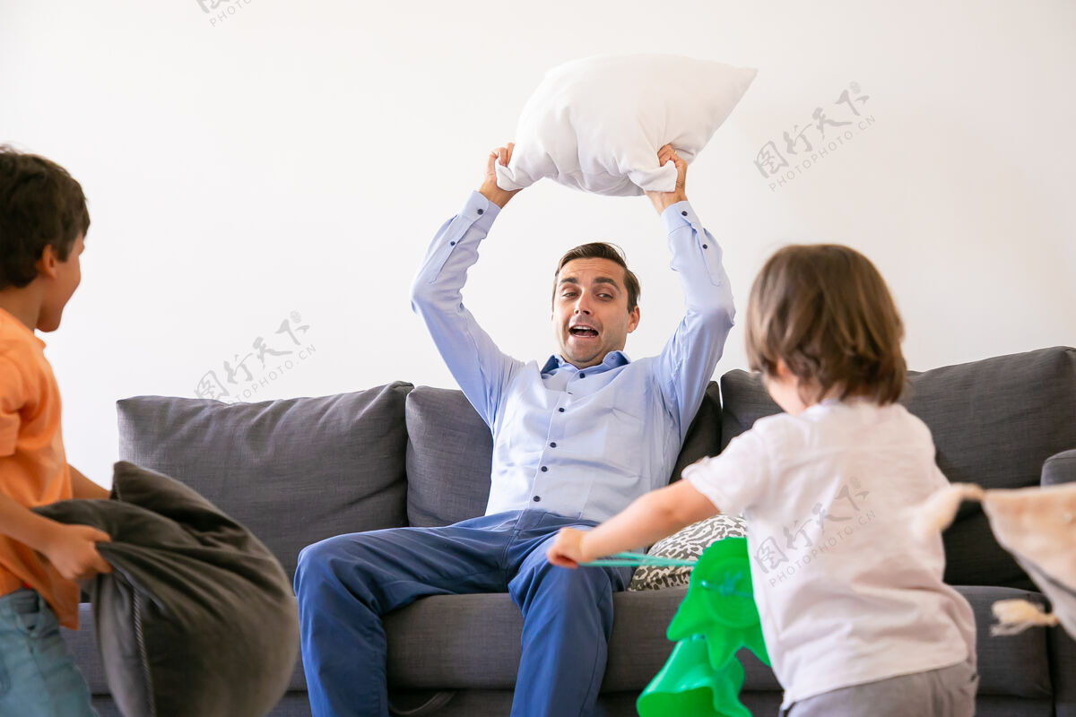 孩子爸爸坐在沙发上 头上抱着枕头快乐的孩子们和爸爸一起玩 一起玩枕头 一起在家里玩童年 节日和游戏活动的概念儿子爸爸女儿