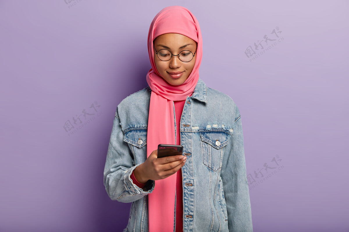 设备穆斯林宗教黑皮女横拍 在现代手机上打信息 戴头巾和牛仔夹克 看通知 上网智能手机站立服装
