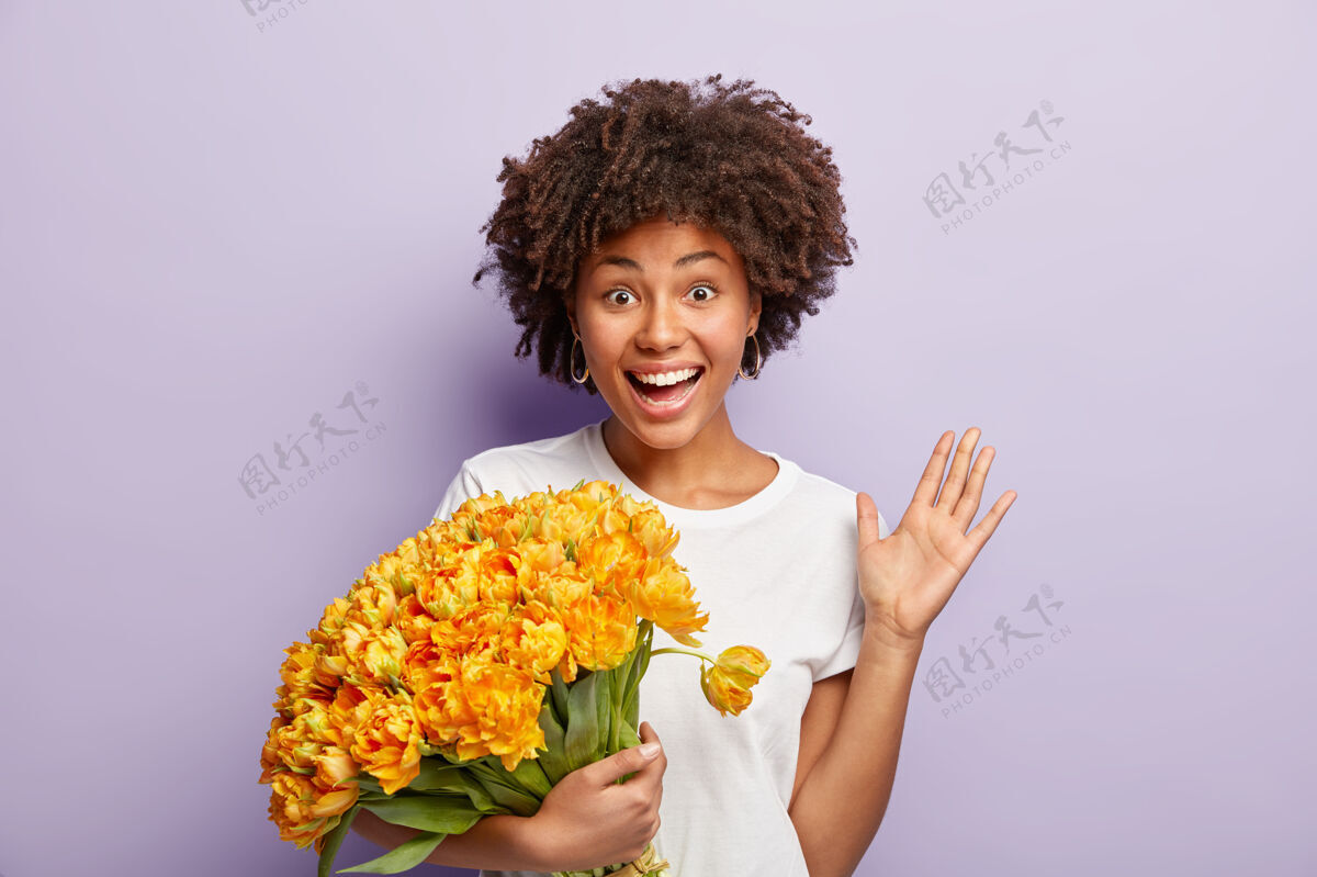 自由空间喜出望外的快乐美女手持芳香的黄色郁金香 挥手致意 向朋友致意 感谢祝贺 留着非洲式发型 穿着白色t恤 紫色墙壁上的模特随意花束女人