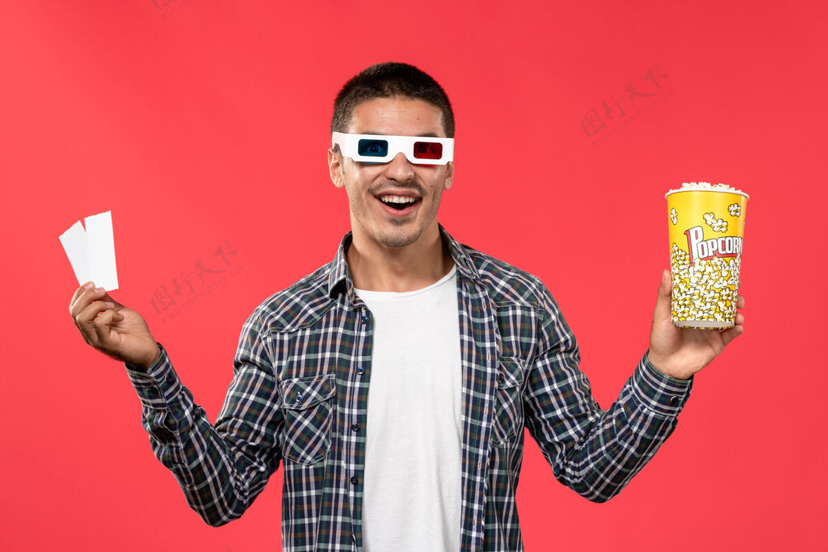 年轻正面图：年轻男子拿着爆米花和票 戴着d型太阳镜 站在浅红色的墙上看男性电影光剧院电影院