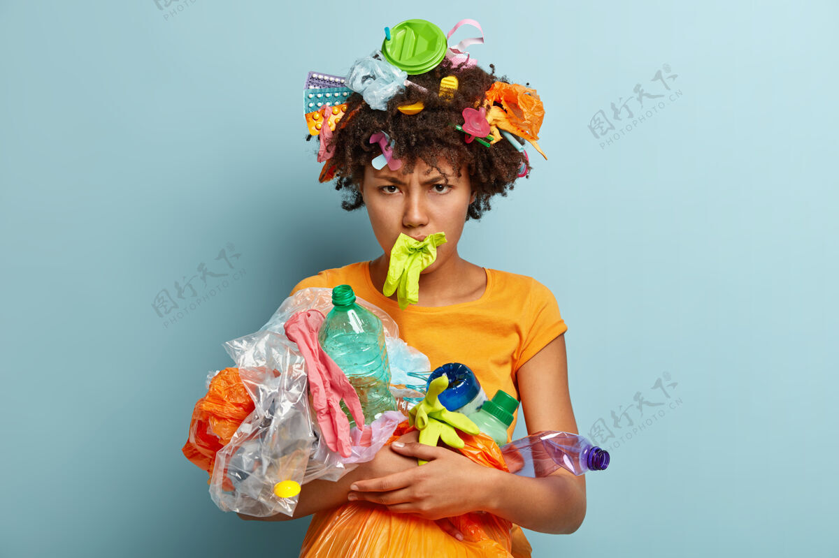 污染郁闷的黑人妇女 清爽的头发 捡垃圾 烦躁的负面表情 清洁被隔离在蓝色墙壁上的环境 整理垃圾 回收 志愿服务的理念清洁环境女人