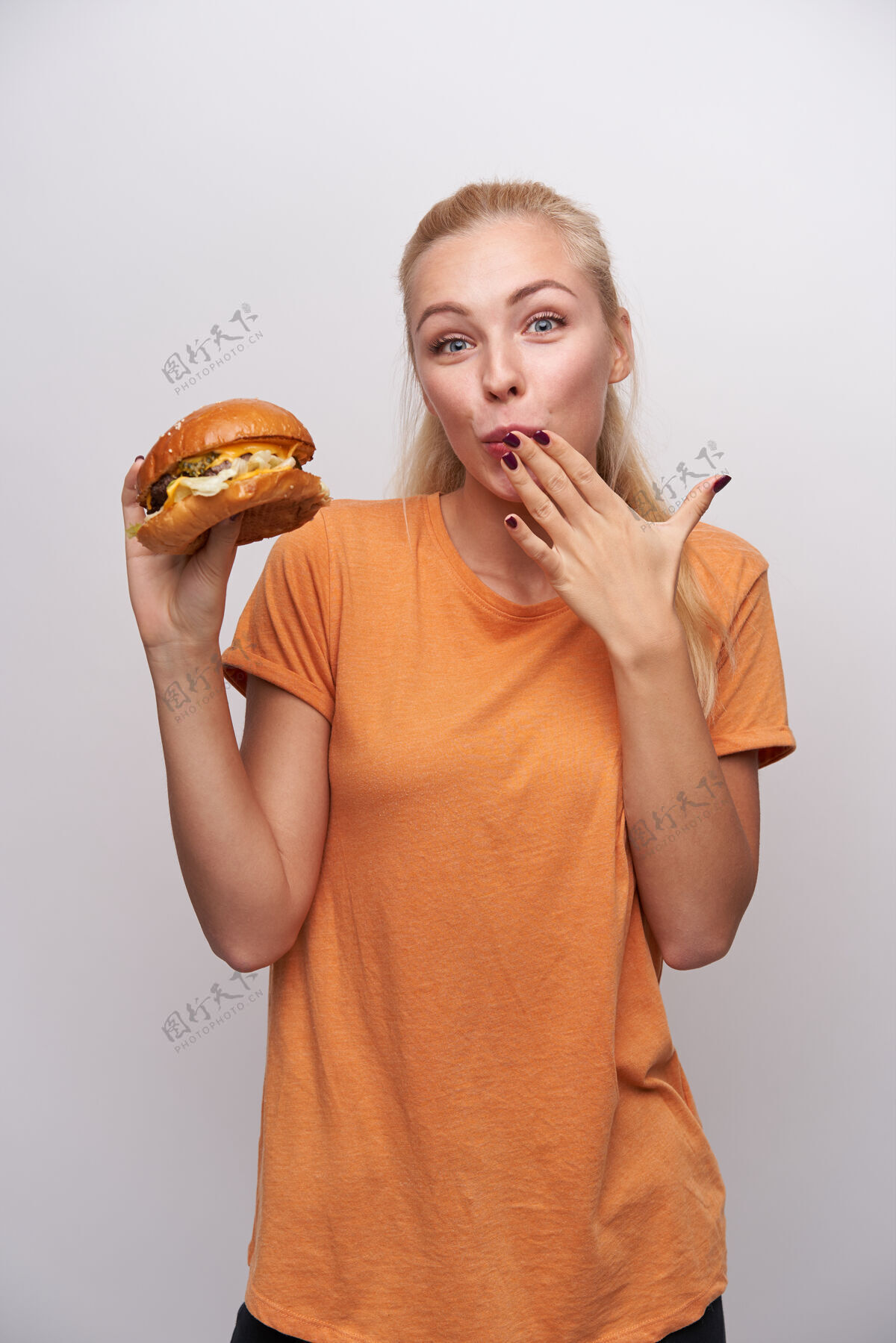 女性快乐的年轻漂亮的金发女郎 随意的发型 正对着镜头 手放在嘴上 一边品尝着新鲜的汉堡 站在白色的背景下食欲饥饿饮食