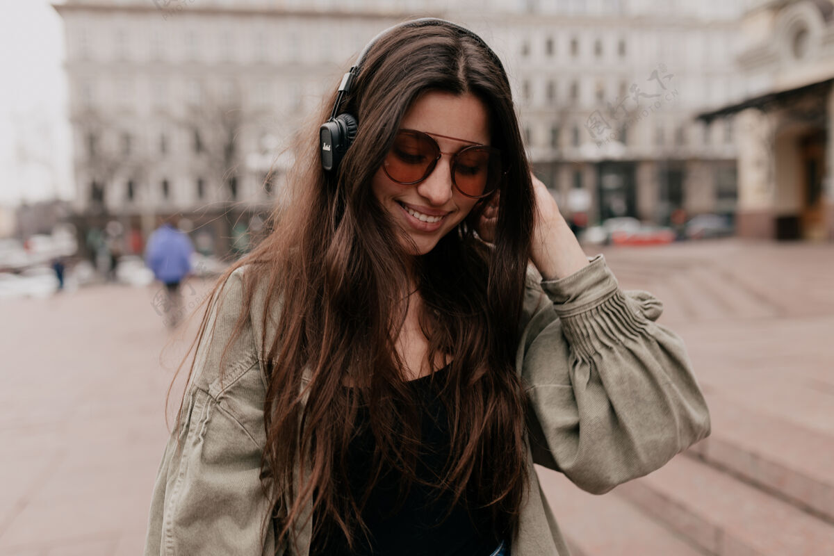 积极快乐微笑的女人在城市里用音乐放松的特写镜头戴着耳机微笑的女模特在周末的户外拍摄年轻都市华丽