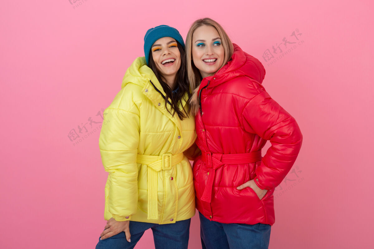 时尚两个迷人的女孩在粉红色的背景下 穿着鲜艳的红色和黄色的彩色冬季羽绒服摆姿势衣服微笑拥抱