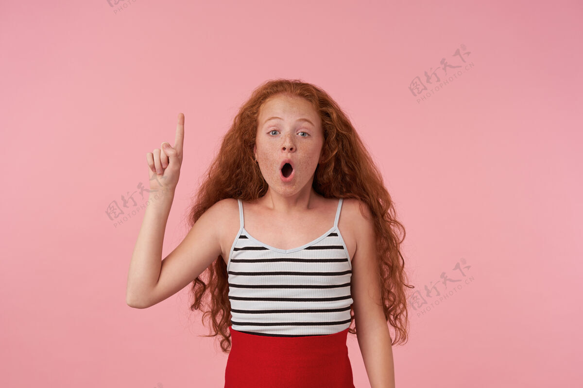 孩子摄影棚拍摄的照片中 一位惊讶的卷发红发女孩穿着优雅的衣服 在粉色背景上摆着休闲发型 张大嘴巴 食指朝上 看着相机室内童年打开