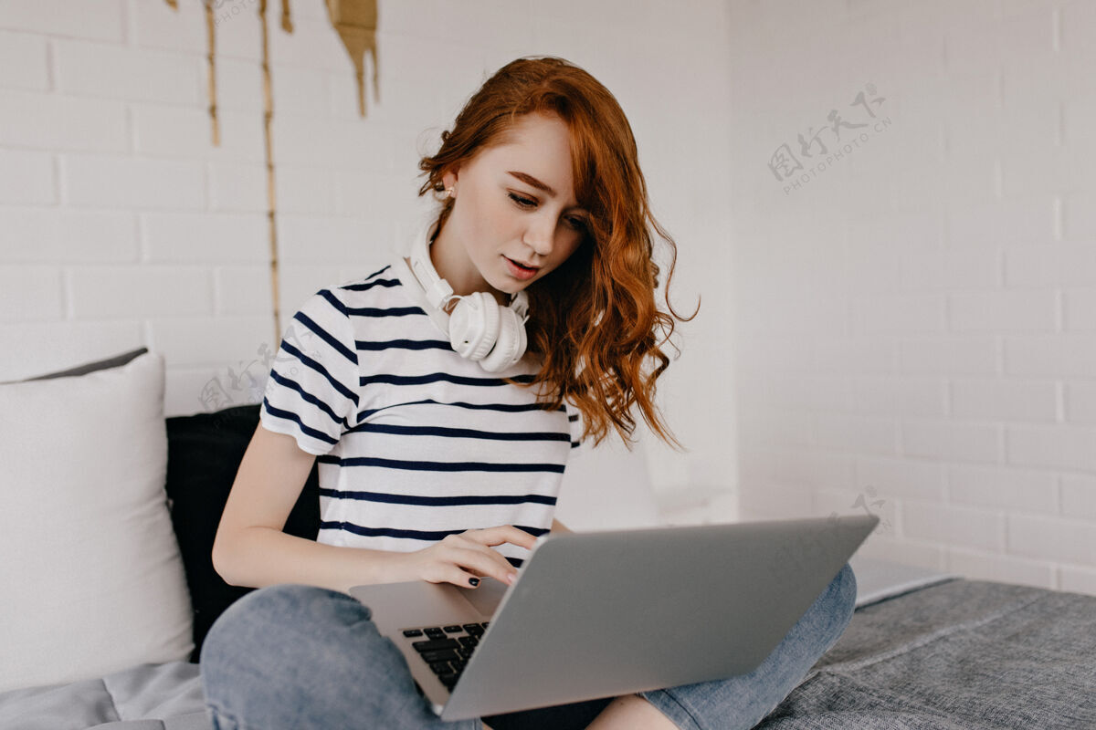 配件专心的卷发女孩坐在床上用键盘打字可爱的姜黄色女士带着笔记本电脑和耳机在家里让人心寒乐趣快乐周末