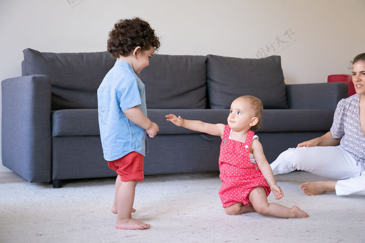 站立可爱的小女孩和混血男孩在地毯上玩耍矮小的妈妈看着孩子们 微笑着卷发的孩子赤脚站在客厅里家庭室内 周末和童年的概念孩子可爱玩耍
